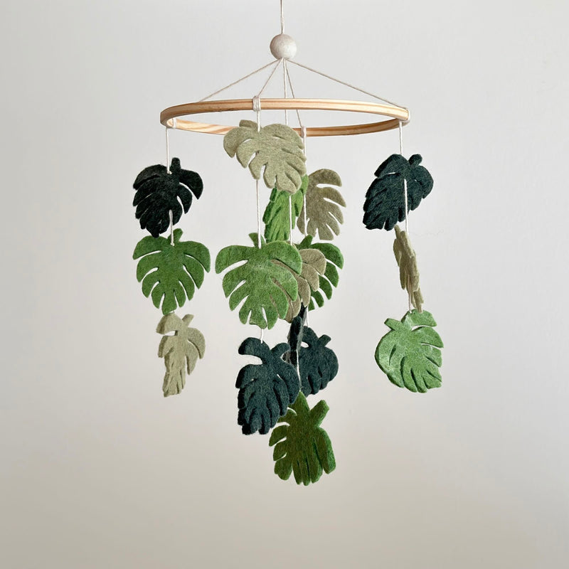 A wooden mobile hangs from a white ceiling, featuring a collection of felt monstera leaves in shades of green. The leaves have darker centers and lighter edges for a realistic appearance, suspended as if floating in the air.