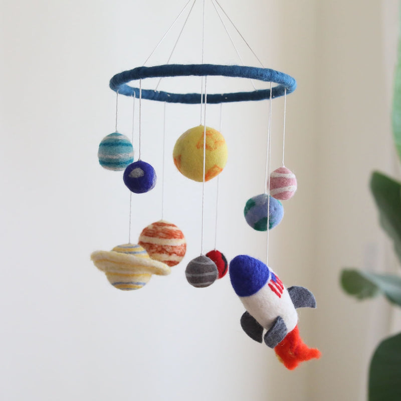A whimsical wool felt baby mobile, featuring planets and a spaceship, hangs from the ceiling in front of a white wall with a plant on the right side. The mobile's central ring showcases vibrant blue, green, orange, and gray tones for the planets, with a red and white spaceship adding a pop of color. Made from soft wool felt, this delightful display adds an air of wonder to any nursery.