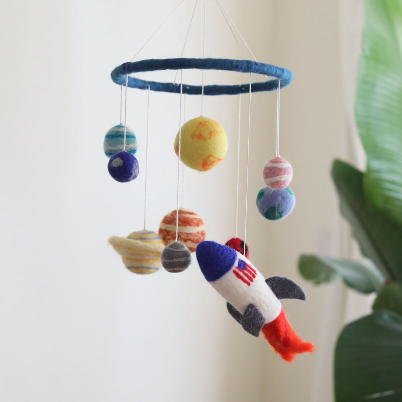A colorful wool felt baby mobile, resembling the solar system, hangs from a string, featuring planets and a spaceship crafted from soft, vibrant wool felt. The mobile is designed for a nursery or crib, adding a touch of magic to a little one's room.