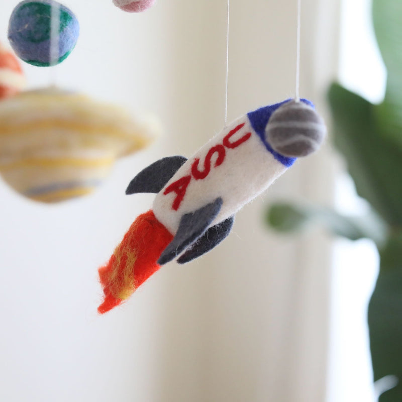A colorful wool felt baby mobile, featuring planets and a spaceship, hangs from a string in a nursery setting. The mobile showcases various shades of blue, green, and brown planets against a predominantly red spaceship with gray details. Made from soft, colorful wool felt, this solar system-themed mobile is perfect for sparking imagination and learning in a child's room.