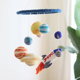 A wool felt baby mobile, Felt Baby Mobile - Solar System, is displayed against a white wall with a plant on the right side. The mobile hangs from the ceiling, featuring a central ring with a blue sky background and planets made from wool felt in shades of yellow, orange, green, and blue. A spaceship designed to look like it's flying through the sky is included as part of the mobile's elements.