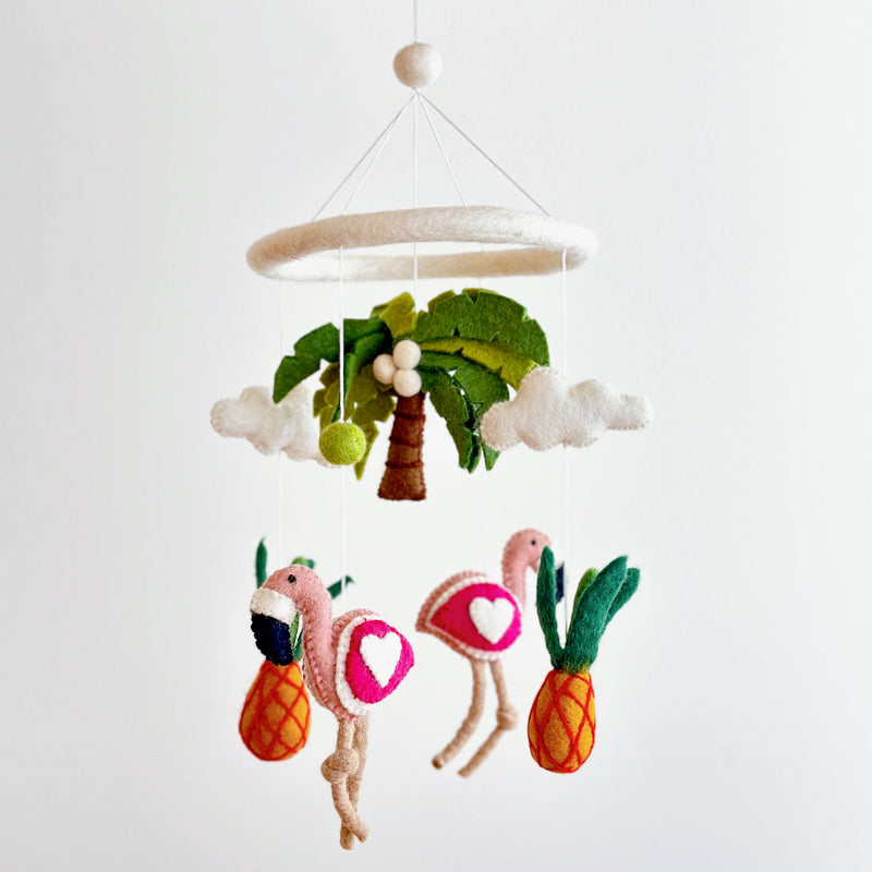 A colorful felt baby mobile, suspended from a white string, features a pineapple, two flamingos, and a palm tree crafted from wool felt. The tropical theme is playful and vibrant, with each element giving off a soft texture. A neutral background allows the colorful elements to stand out.
