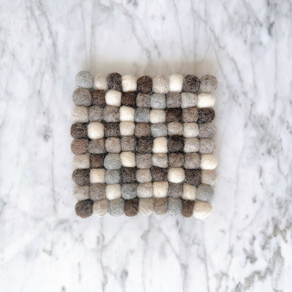 A gray wool felt ball square cup coaster is presented in a top-down view on a white marble surface. This handcrafted felt coaster, part of a set, is meticulously made from 100% organic lamb wool felt. Its surface features numerous gray felt balls, neatly hand-stitched together, creating a unique, tactile texture. Ethically made by Nepalese female artisans, it supports fair-trade craftsmanship.