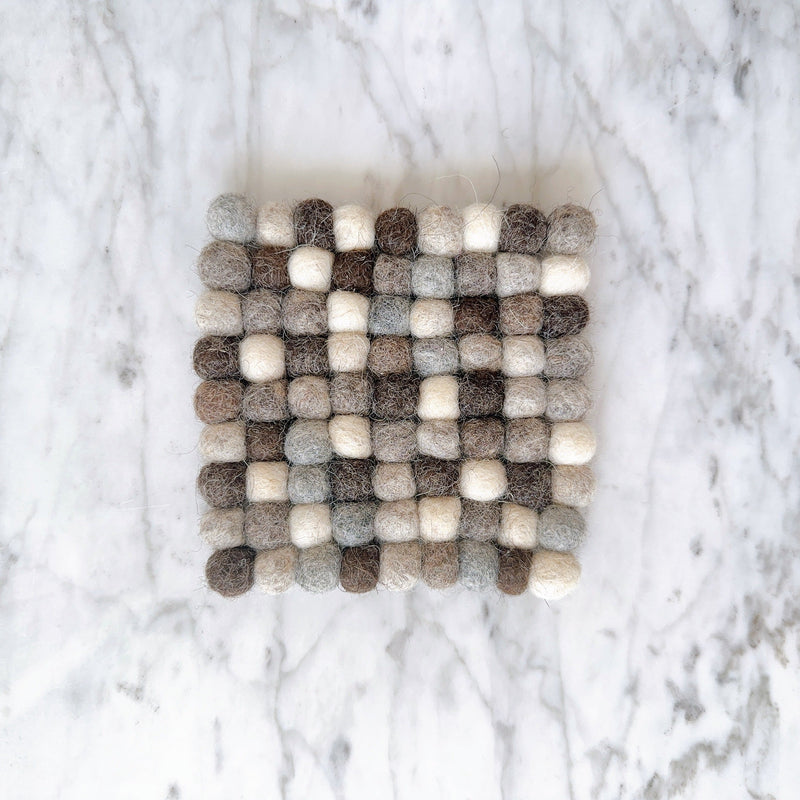 A gray wool felt ball square cup coaster is presented in a top-down view on a white marble surface. This handcrafted felt coaster, part of a set, is meticulously made from 100% organic lamb wool felt. Its surface features numerous gray felt balls, neatly hand-stitched together, creating a unique, tactile texture. Ethically made by Nepalese female artisans, it supports fair-trade craftsmanship.