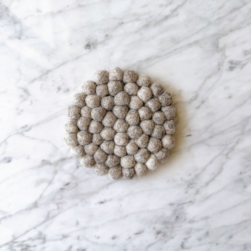 A single, round gray felt ball coaster is displayed on a white marble surface with subtle gray veining. This handcrafted felt coaster is composed of numerous tightly packed, hand-stitched felt gray balls made from 100% organic lamb wool. Its minimalist design, ethically handmade by Nepalese female artisans, evokes calm and understated elegance.
