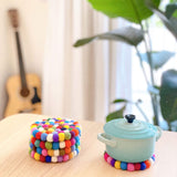 Five vibrant felt ball rainbow round cup coasters rest on a light wooden table. Four handmade felt coaster is stacked on one side, showcasing its thickness and the colorful, hand-stitched felt balls crafted from 100% organic lamb wool. The other coaster holds a light blue cooking mini pot, demonstrating its heat resistance. A blurred acoustic guitar and a green plant add to the cozy home decor.