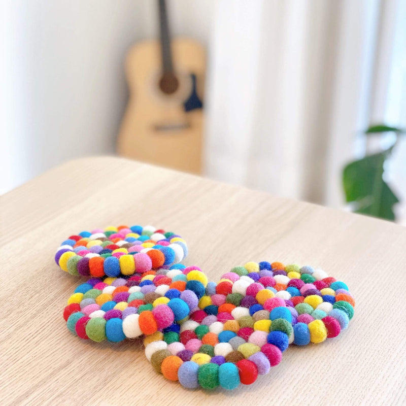 Four round wool felt rainbow cup coasters are cheerfully arranged on a light wooden table, with a guitar and leafy plant softly blurred in the background. Each handcrafted felt coaster is made from small, hand-stitched felt balls of 100% organic lamb wool, displaying vibrant, blended colors of white, yellow, orange, pink, red, purple, blue, and green. These unique, ethically handmade felt coasters add an inviting, artistic touch to any space.