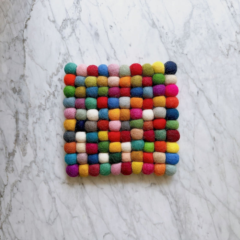 A vibrant square felt ball rainbow coaster rests on a cool marble surface. This handcrafted felt coaster, part of a set, is made from 100% organic lamb wool, with numerous hand-stitched felt balls forming a square pattern. It measures 4 x 4 inches and displays cheerful rainbow hues, showcasing the fair-trade craftsmanship of Nepalese female artisans.