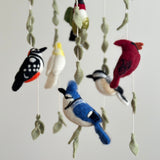 A vibrant felt bird baby mobile, part of the 'Wings of America' collection, is displayed against a light grey background. It features five distinct felt birds: a red cardinal, a green and white hummingbird, a blue jay, a black and white chickadee, and a yellow and white parrot. Each bird hangs at varying heights from a wooden frame, interspersed with decorative felt leaves, creating a charming, nature-inspired nursery decor.