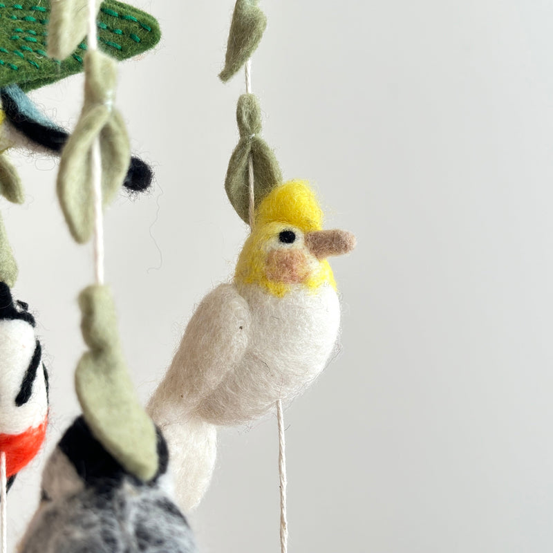 A charming felt bird baby mobile is shown in a close-up against a plain white background, highlighting its intricate details. Several handcrafted felt birds and decorative leaves hang from a natural wooden frame by white strings. One prominent bird features a yellow head and white body, while the felt leaves display visible stitching.