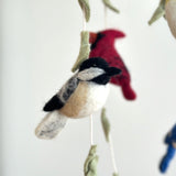 A colorful wool felt bird baby mobile hangs against a clean white background. It features a whimsical avian theme with various birds crafted from soft wool felt on a wooden frame. Prominent among them are a chickadee with a black cap and pale beige body, a red cardinal, and green leaves, alongside a glimpse of a blue jay.