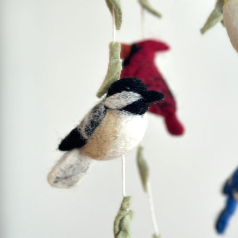 A colorful wool felt bird baby mobile hangs against a clean white background. It features a whimsical avian theme with various birds crafted from soft wool felt on a wooden frame. Prominent among them are a chickadee with a black cap and pale beige body, a red cardinal, and green leaves, alongside a glimpse of a blue jay.