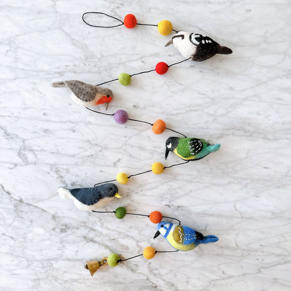 A charming wool felt bird garland is displayed against a bright white marble background. It features five delightful 3D felt birds: a woodpecker with a white belly and black back, a robin with a grey back and orange chest, a blue tit with blue wings and yellow chest, a nuthatch with a white chest and dark grey wings, and a great tit with a green back and yellow chest. Colorful felt balls in red, orange, yellow, green, and purple separate the birds, with a copper bell at the end.