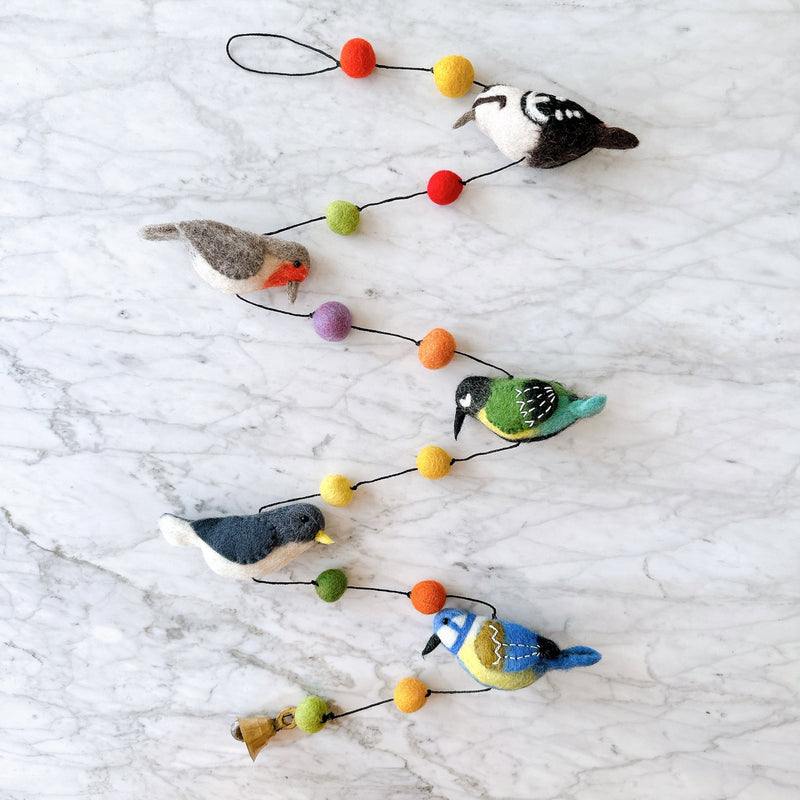 A charming wool felt bird garland is displayed against a bright white marble background. It features five delightful 3D felt birds: a woodpecker with a white belly and black back, a robin with a grey back and orange chest, a blue tit with blue wings and yellow chest, a nuthatch with a white chest and dark grey wings, and a great tit with a green back and yellow chest. Colorful felt balls in red, orange, yellow, green, and purple separate the birds, with a copper bell at the end.