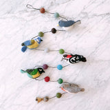 A wool felt bird garland is displayed on a white marble surface. It features five handcrafted felt birds: a woodpecker, robin, blue tit, nuthatch, and great tit. The birds are connected by a string with colorful felt balls and finished with a small copper bell.