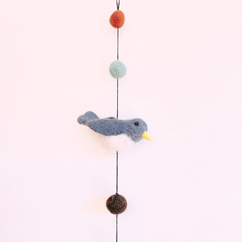 A colorful felt bird garland hangs vertically against a plain white background. It showcases a detailed 3D needle-felted  Nuthatch bird with a white belly, yellow beak, and small black eye. Above the bird are light green and orange wool felt balls, with a brown felt ball below. The charming wool felt garland is suspended by a black string, ending with a small copper bell.