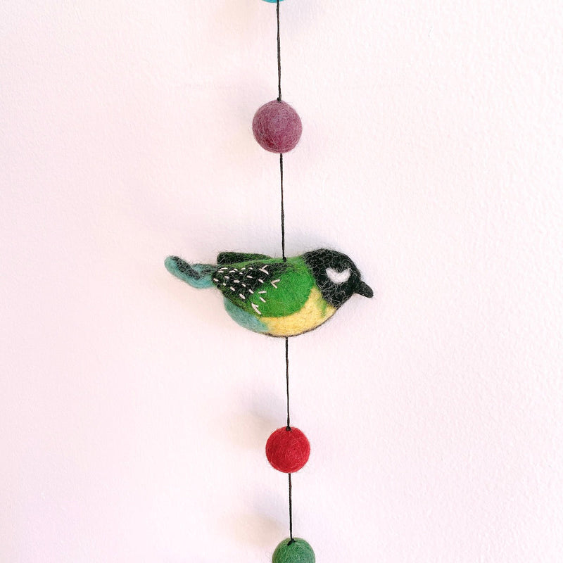 A vibrant wool felt bird garland hangs against a plain white wall. A close-up highlights a cheerful green Great Tit bird, featuring a black head, green wings with white accents, and a yellow belly. It is suspended from a black string, with a purple felt ball above and red and green felt balls below, all crafted from 100% wool felt.