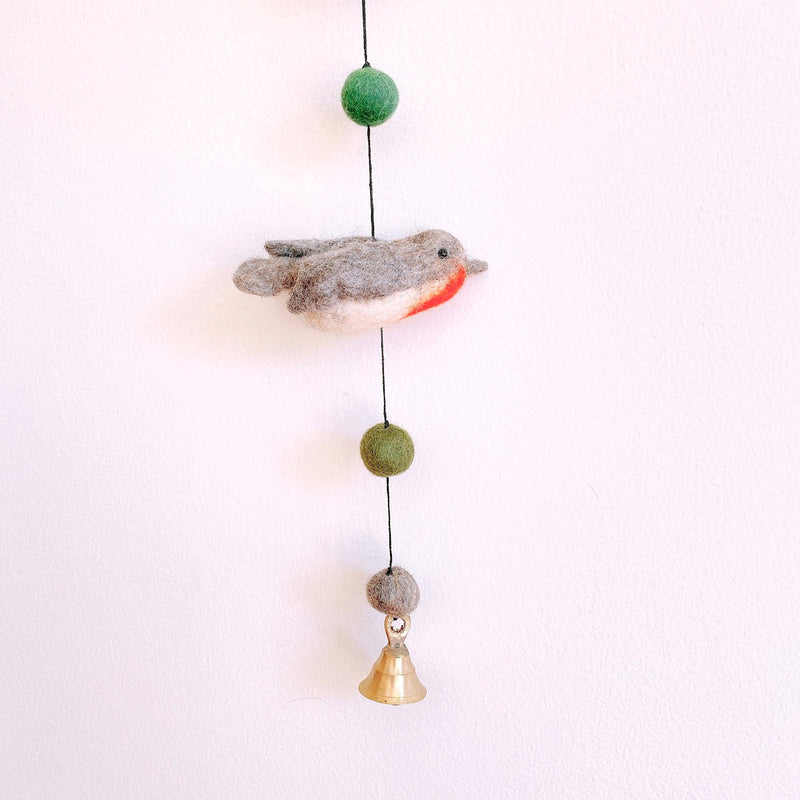 A handcrafted wool felt bird garland is displayed against a simple white backdrop. This charming felt garland features multiple birds, including a gray, white, and red Robin. It also includes green and gray wool felt balls strung along a black cord, finished with a copper bell at the bottom, adding a whimsical touch.