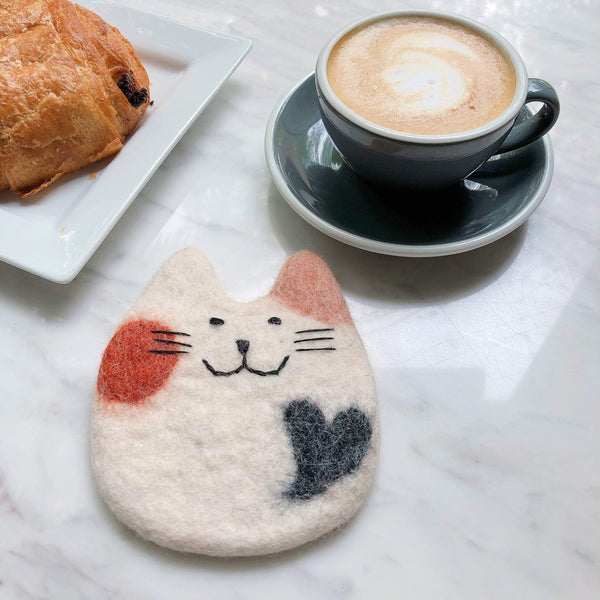 A unique, handmade felt calico cat coaster, part of a set, rests on a white marble table alongside a coffee cup and pastry. This sheep wool felt coaster is shaped like a charming calico cat, primarily white with a red spot, a pink ear, and a black heart-shaped spot. Black needle-felted details form its smile, nose, and whiskers, showcasing the ethical craftsmanship by Nepalese female artisans.