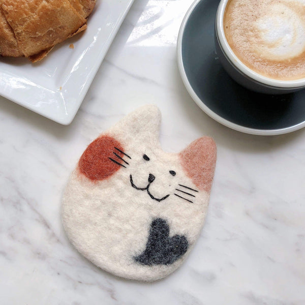 A charming needle felted calico cat coaster, part of a Felt Cat & Dog Coaster set, rests on a white marble surface next to a pastry and a gray cup. This handcrafted wool felt coaster features a smiling cat face with soft, rounded ears, detailed with black and brown markings on a white base. Ethically handmade, this unique coaster brings a cozy, playful vibe.