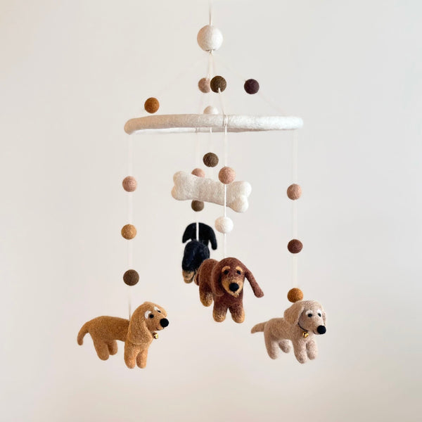 A Felt Dachshund dog baby mobile hangs against a neutral, off-white backdrop. It features four soft felt dachshunds in cream, khaki, brown, and black, suspended from a white felt frame. A white felt bone is centered above the dogs, creating a comforting aesthetic.