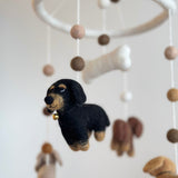 A charming felt dachshund dog baby mobile is presented in a close-up against a soft white background, creating a calm atmosphere. The mobile features a white felt frame with a white felt bone at its center. Visible dachshunds include one black and tan dog in front, and others in tan and brown.