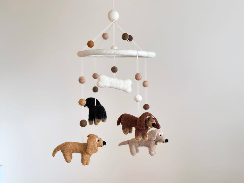 A felt Dachshund baby mobile is shown against a muted off-white background. Four felt dachshunds in cream, khaki, brown, and black hang from a white felt frame, arranged in a circular pattern. A white felt bone hangs playfully in the center.