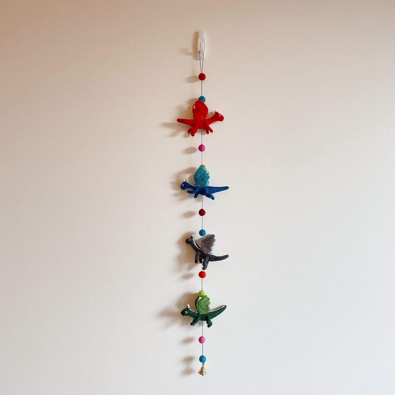 A colorful felt dragon garland is displayed against a plain off-white wall. The garland features four 3D felt pterosaurs in red, blue, gray, and green. They are strung along with colorful felt balls, and a small copper bell hangs at the bottom.
