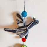 A section of a gray felt dragon garland is displayed against a plain white background. The prominent gray wool felt pterosaur is a 3D flying dragon, detailed with white stripes on its wings and body, giving it a soft, textured appearance. Small white horns and simple black and white eyes add whimsical charm. Connected by a thin black string, a copper bell is also visible on this handcrafted nursery garland.