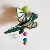 A green wool felt dragon, part of a Felt Dragon Garland, hangs suspended from a thin black string against a bright white background. The three-dimensional dragon is crafted from soft green felt with white accents on its wings and body. Below the dragon, a copper bell is attached to the string.