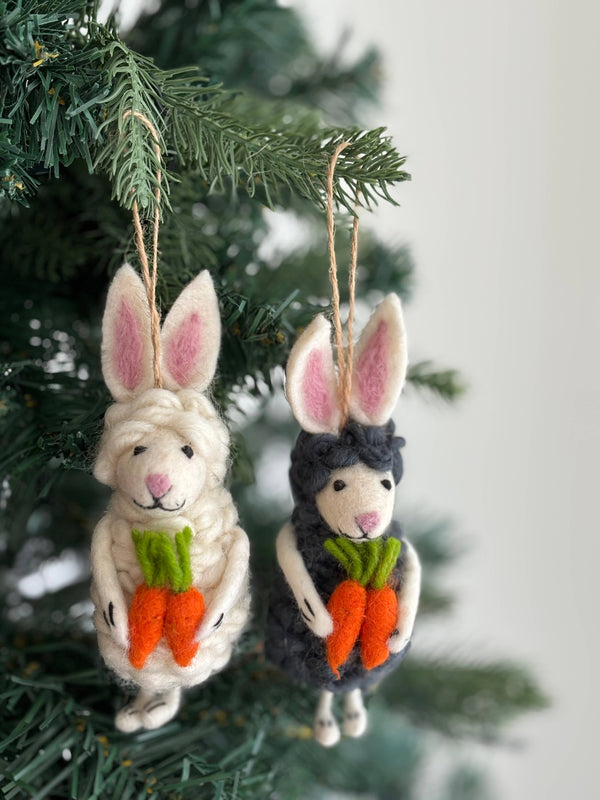 Two felt sheep bunny ornaments are charmingly displayed on a green Christmas tree branch. These fluffy felt ornaments include a white one with pink-lined ears and a black one with dark, pink-lined ears. Both unique Christmas ornaments hold small orange carrots with green tops and hang by jute strings.