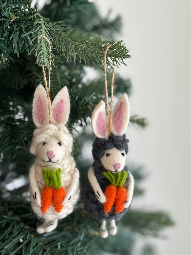 Two felt sheep bunny ornaments are charmingly displayed on a green Christmas tree branch. These fluffy felt ornaments include a white one with pink-lined ears and a black one with dark, pink-lined ears. Both unique Christmas ornaments hold small orange carrots with green tops and hang by jute strings.