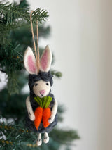 The wool felt Easter bunny sheep ornament hangs from a jute string on a tree, against a softly blurred evergreen background. This playful needle felted sheep features a grey wool body with white feet, a light pink nose, and black eyes. It is adorned with white bunny ears with pink insides and holds two orange carrots with green tops.