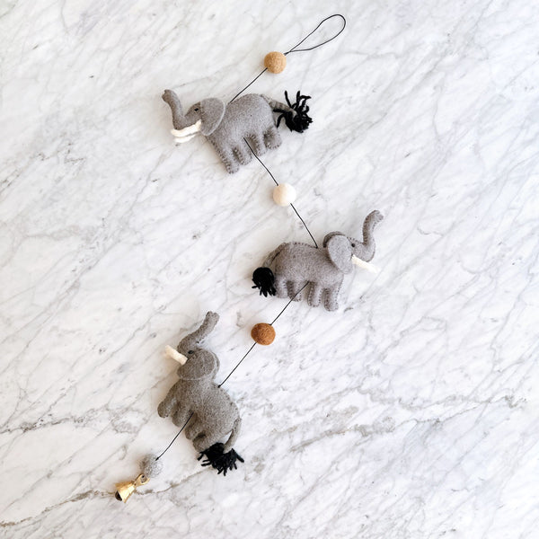 A whimsical felt elephant garland rests on a bright, clean marble surface. The garland features three gray felt elephants, each with a white felt tusk and a small black yarn tail. They are strung on a thin black string, interspersed with tan, white, and brown felt balls, with a copper bell dangling at one end.
