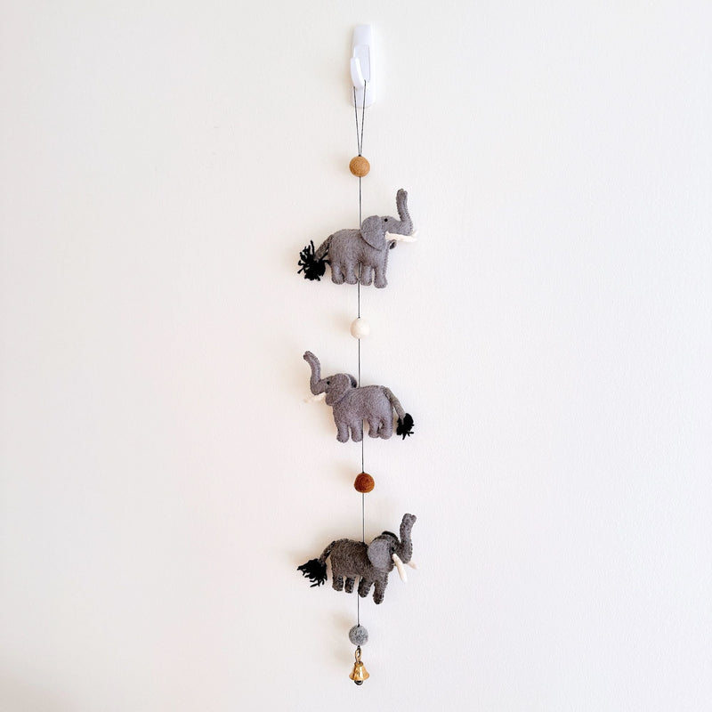 A felt elephant garland is suspended from a white hook against a plain white wall. The garland features three gray felt elephants, each detailed with white tusks and black yarn tails. Round felted balls in brown and white separate the elephants. A small copper bell is attached at the bottom.