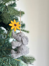 A gray wool felt elephant ornament, with a textured body and floppy ears, hangs from the green branches of a Christmas tree. The adorable elephant holds a yellow flower with a brown center and green stem in its trunk. A jute string allows this charming felt Christmas ornament to be suspended, creating a cozy and festive atmosphere.