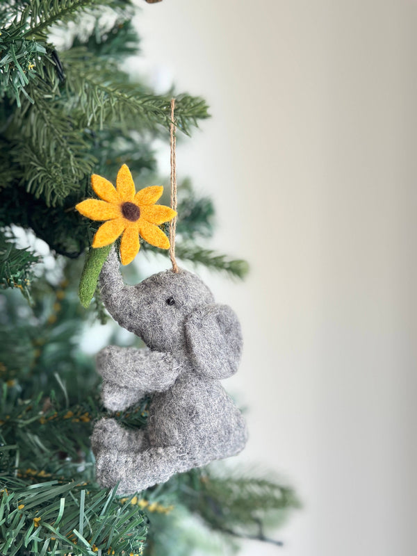 A gray wool felt elephant ornament, with a textured body and floppy ears, hangs from the green branches of a Christmas tree. The adorable elephant holds a yellow flower with a brown center and green stem in its trunk. A jute string allows this charming felt Christmas ornament to be suspended, creating a cozy and festive atmosphere.