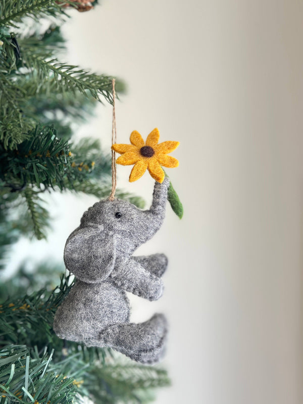 A gray wool felt elephant ornament, holding a yellow flower, is displayed hanging on the green branches of a Christmas tree. The textured elephant features small black eyes and large, distinct ears. Its trunk holds a vibrant yellow felt flower with a brown center and green stem. A jute string is attached for graceful suspension.