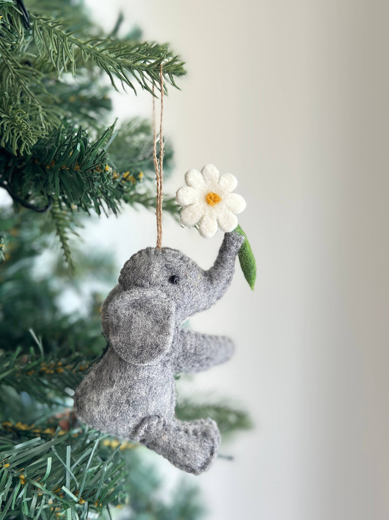 A handcrafted felt elephant with flower ornament hangs gracefully from a green Christmas tree branch. This soft gray felt elephant ornament features a black eye, meticulously shaped ears, and a trunk playfully holding a white felt flower with a yellow center and green stem. A jute string allows it to hang beautifully.