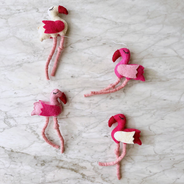 A vibrant felt flamingo garland is displayed on a bright white marble surface. This cheerful wool felt garland, approximately 4 feet long, features four needle-felted flamingos in varied pink and white hues, some with whimsical closed eyes and thin legs. A charming copper bell dangles from one end.