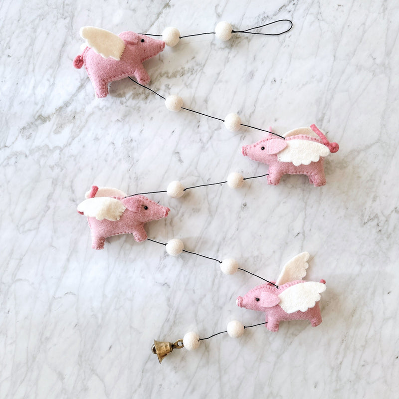 A whimsical wool felt flying pig garland is displayed against a light marble background. The charming felt garland features four pink pig figures, each adorned with white felt wings, strung with white felt balls on a thin black cord. Each pig is detailed with small black eyes, pink snouts, and curly tails.