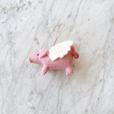 A single pink felt flying pig, a whimsical component of a felt flying pig garland, rests on a bright, clean marble surface. The handmade pink wool felt piggy bird features small button eyes and white felt wings. Its detailed stitching gives it a textured appearance, and a small string is attached for easy hanging.