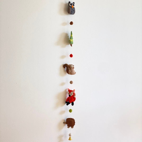 A colorful wool felt forest animal garland hangs vertically against a plain white wall. It features needle-felted 3D figures, including a gray and white owl, a green tree, a tan squirrel, an orange and white fox, and a brown hedgehog. Small, colorful felt balls separate the figures, with a copper bell providing an elegant finish at the bottom.