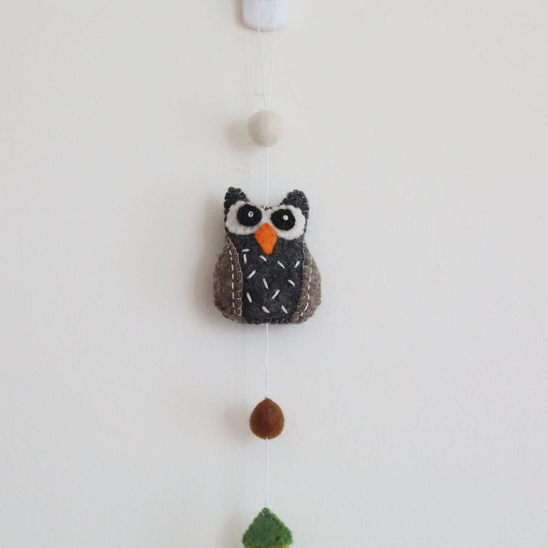 A wool felt forest animal garland is displayed against a plain white wall, suspended from a subtle white hook. The handmade garland features a detailed 3D owl with a gray body, brown accents, white stitching, and an orange beak. Above the owl is a white felt ball, while a green felt tree hangs below, connected by a thin string. A small copper bell completes the natural, handcrafted design.
