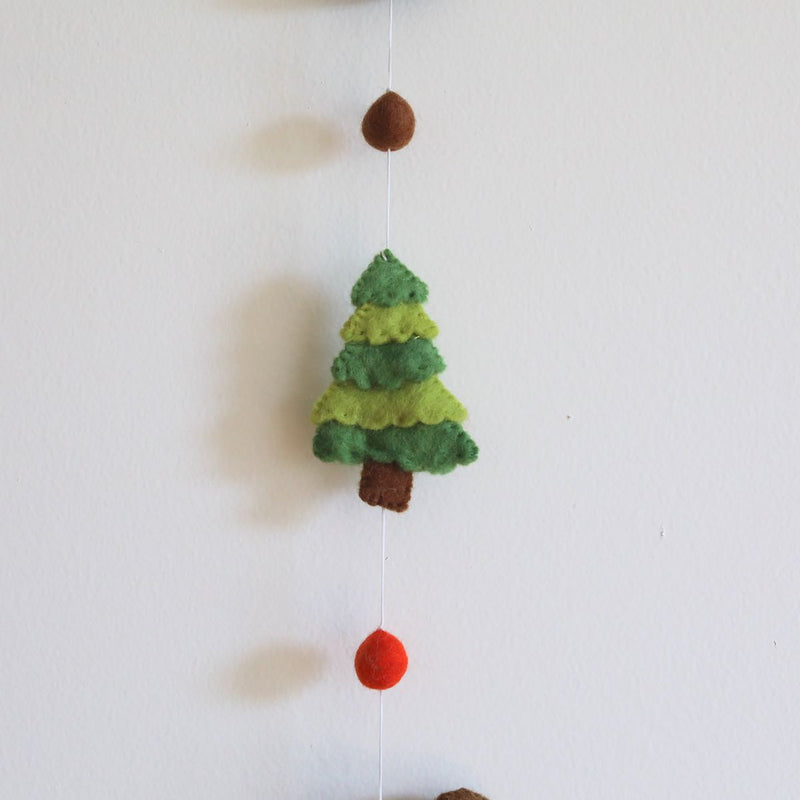 A charming green felt tree, part of a felt forest animal garland, is displayed against a plain white wall. Crafted from soft, textured 100% wool felt, this needle-felted tree features three 3D layers in varying green shades, supported by a brown trunk. A thin white string connects it to a small brown felt ball above and a red felt ball with another forest animal below, creating a whimsical forest scene.