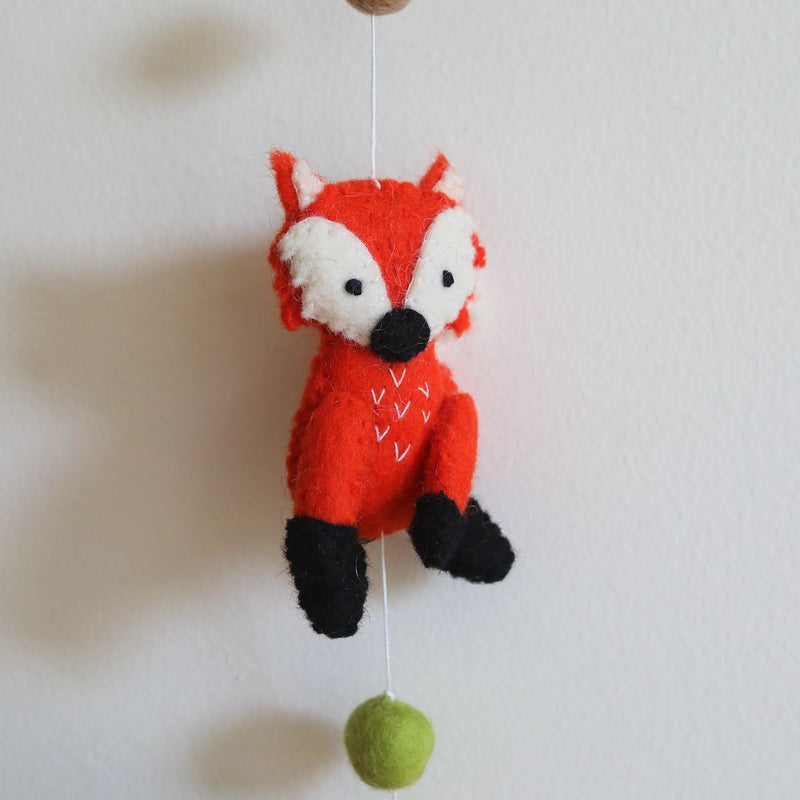 A vibrant orange felt fox, part of a Felt Forest Animal Garland, is suspended against a light, neutral background. This 3D-designed, handmade wool felt fox features white and black accents with small stitched details on its chest, showcasing its textured, needle-felted appearance. A small green felt ball hangs below the fox, completing the cheerful design.