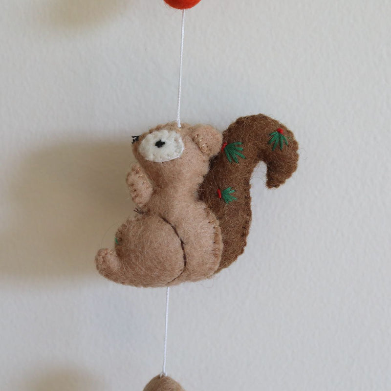 A light brown, 3D needle-felted squirrel from a felt forest animal garland hangs against a plain white wall. This 100% wool squirrel features a darker brown tail adorned with small green and red embroidered details. It is suspended by a thin white string, with a red felt element above and a small brown felt ball below, suggesting the garland extends to a copper bell.