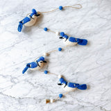 A Blue Jay felt garland is flat laid on a white marble surface. It features four meticulously crafted blue and white blue jays, each with fluffy fur atop their heads. They are evenly spaced along a thin, rustic twine, interspersed with small alternating white and blue felt balls, creating a delicate and charming display.