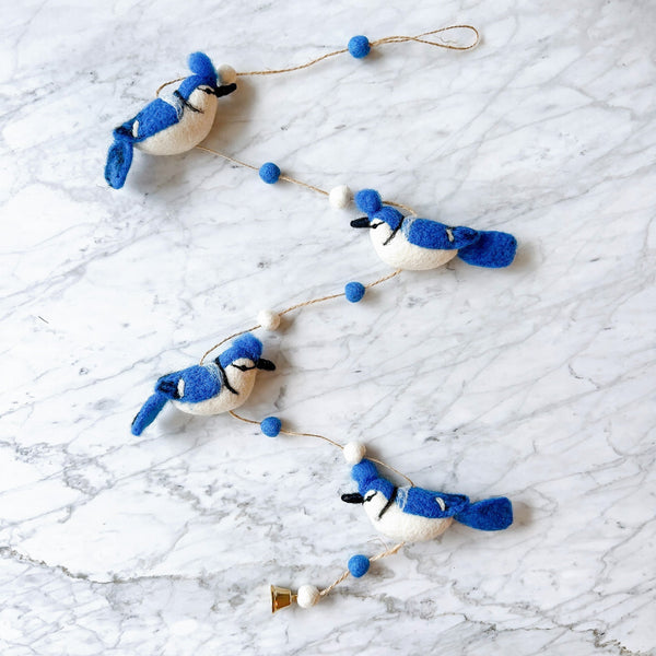 A Blue Jay felt garland is flat laid on a white marble surface. It features four meticulously crafted blue and white blue jays, each with fluffy fur atop their heads. They are evenly spaced along a thin, rustic twine, interspersed with small alternating white and blue felt balls, creating a delicate and charming display.