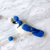 The vibrant felt blue jay garland, featuring four meticulously crafted blue jays, is elegantly displayed on a textured white surface. Each blue jay boasts striking blue tones on its head, wings, and tail, complemented by a soft white body and fluffy fur. Rustic twine with playful wool balls connects the charming birds, creating a bright and airy atmosphere.